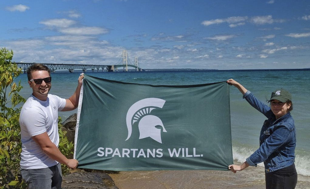 Two people holding Spartans Will flag on the shoreline